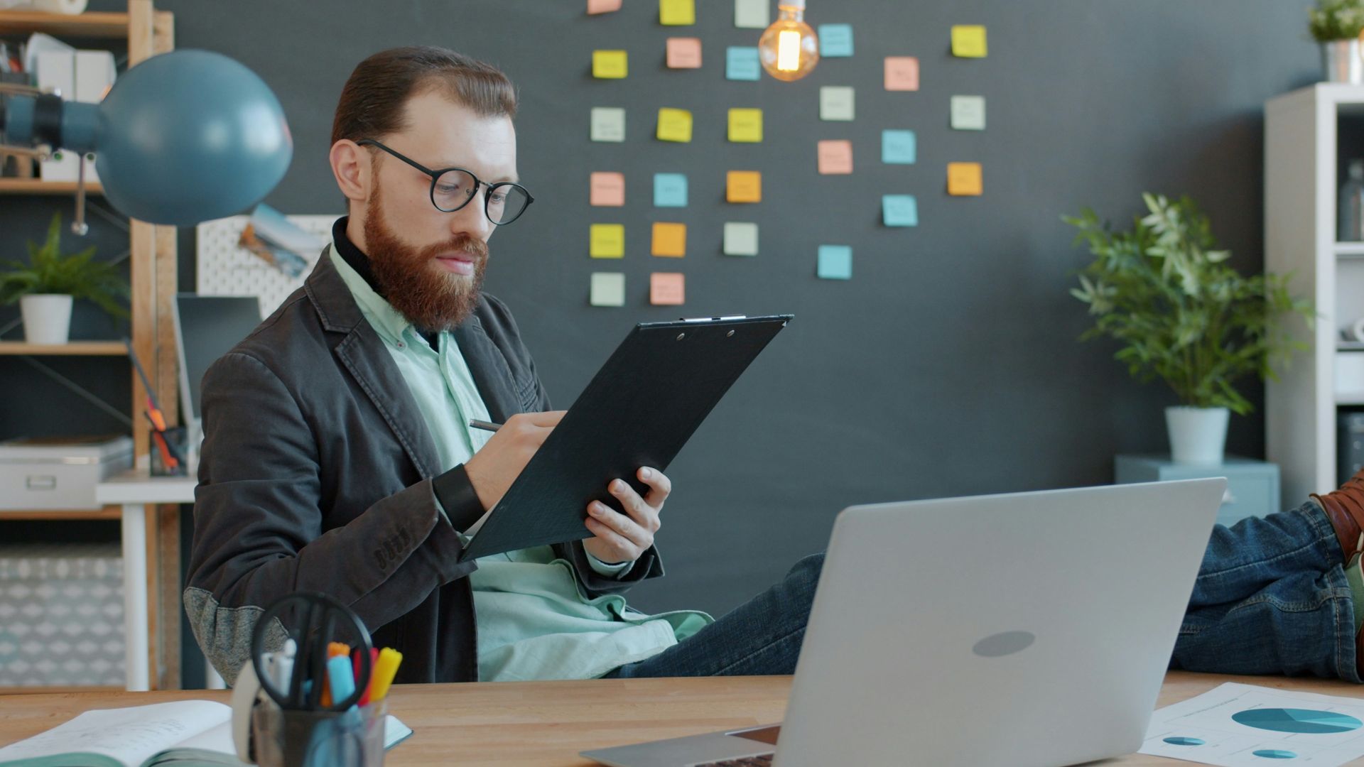 Man with beard working at desk with laptop and papers.