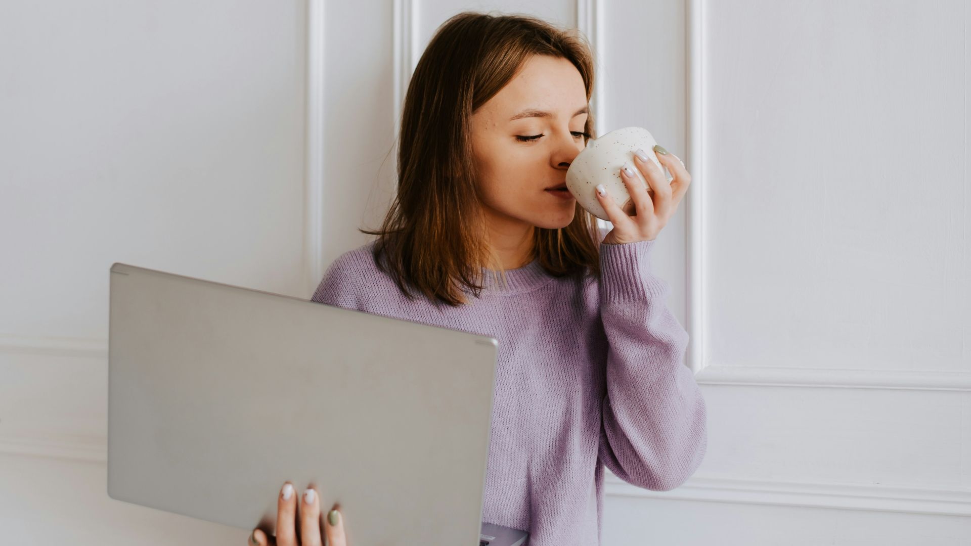 a woman drinking from a cup while holding a laptop
