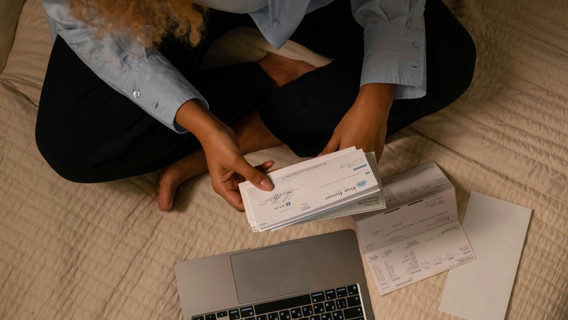 Overhead view of woman organizing finances on bed with laptop and checks.