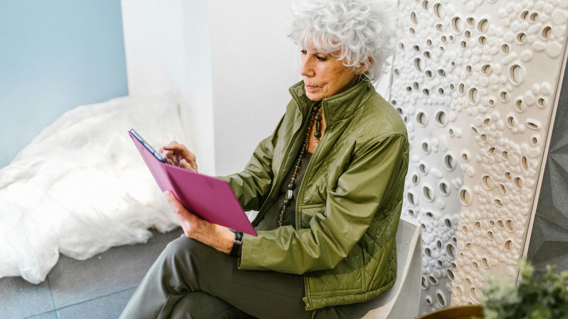 Elderly woman in green jacket using a tablet indoors, showcasing technology use in modern lifestyles.