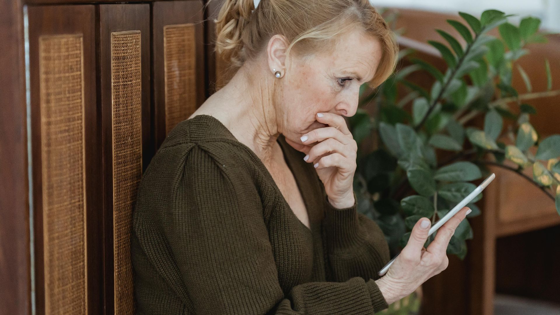 Side view of surprised elderly female with cellphone covering mouth against cabinet and plant at home