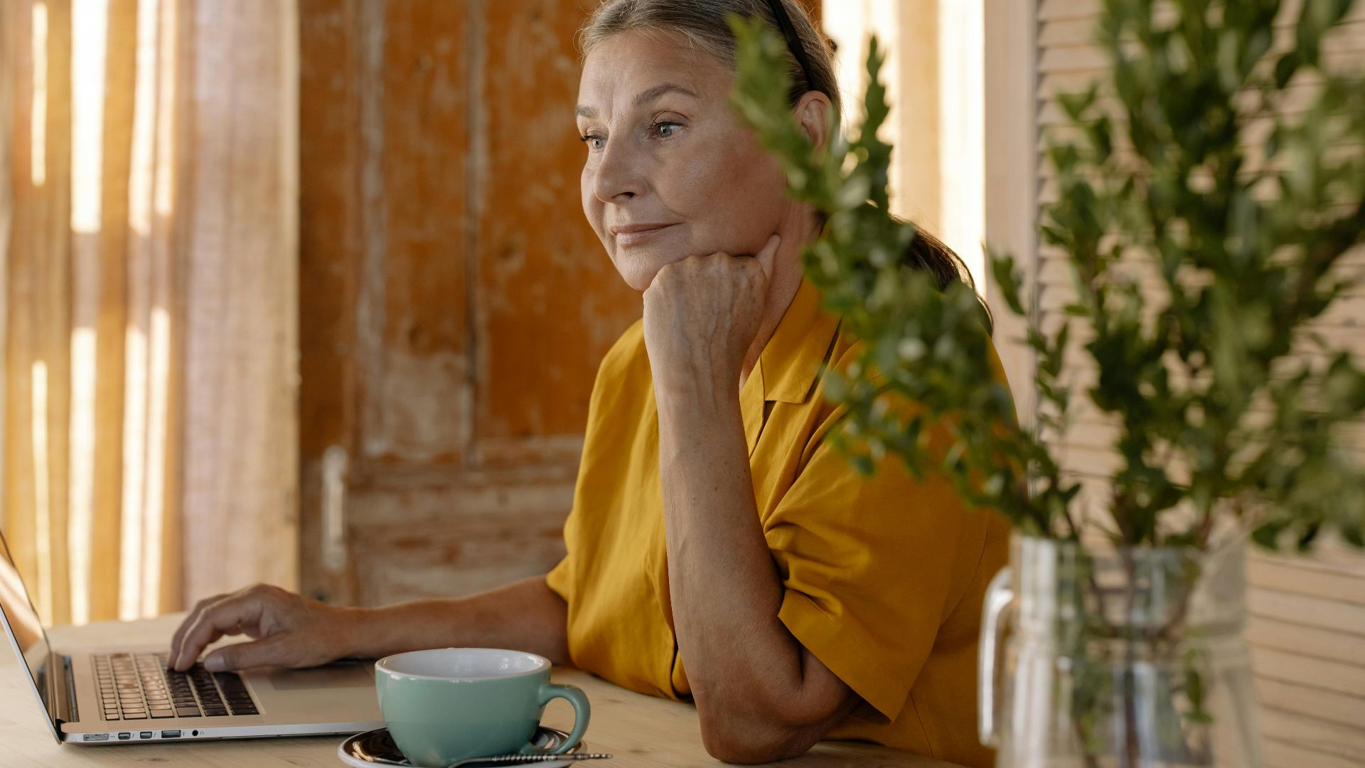 Elderly woman sitting at a table using a laptop, relaxing in a cozy home setting.