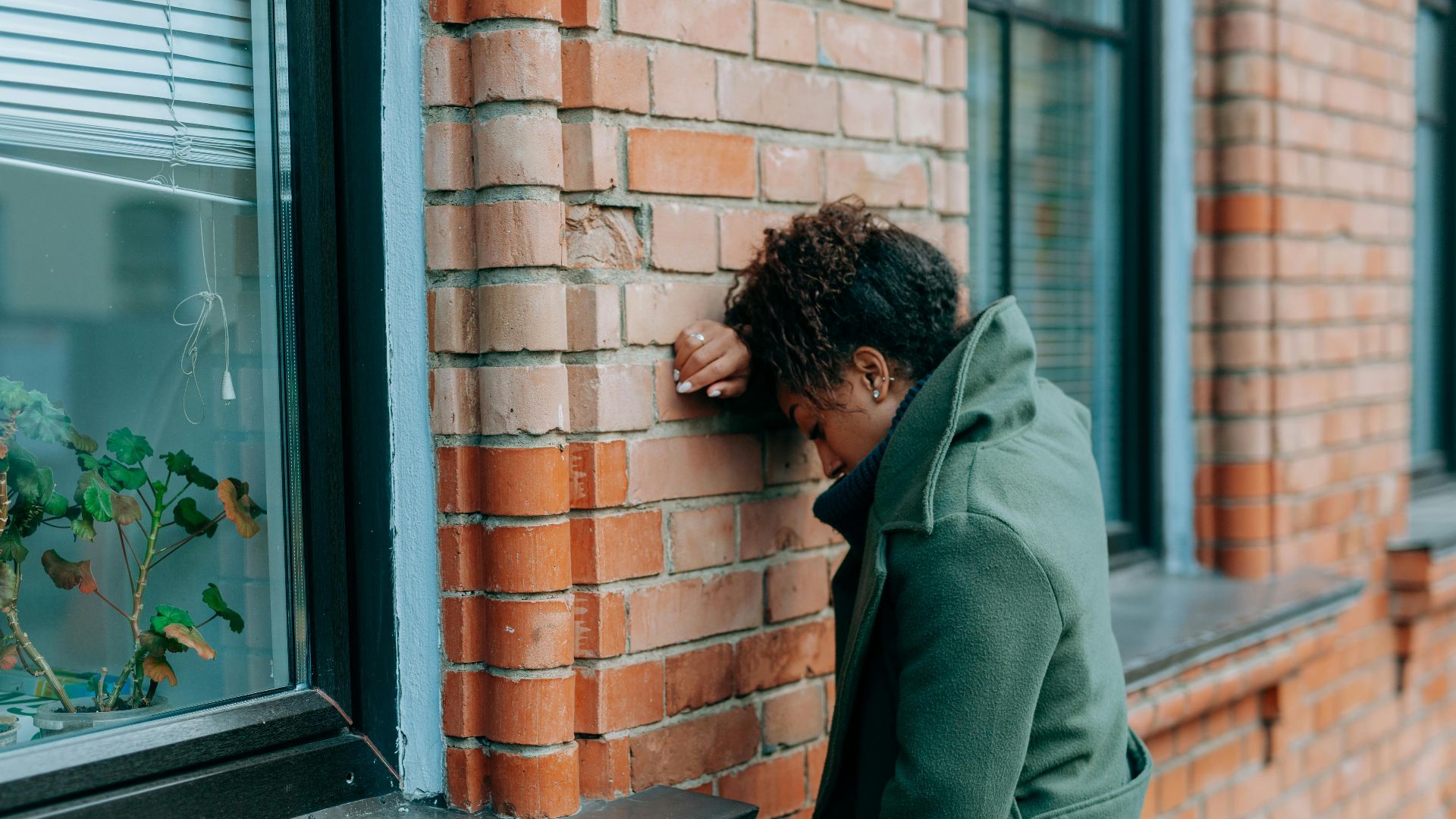 A woman in a green coat leans against a brick wall, reflecting deeply outdoors.