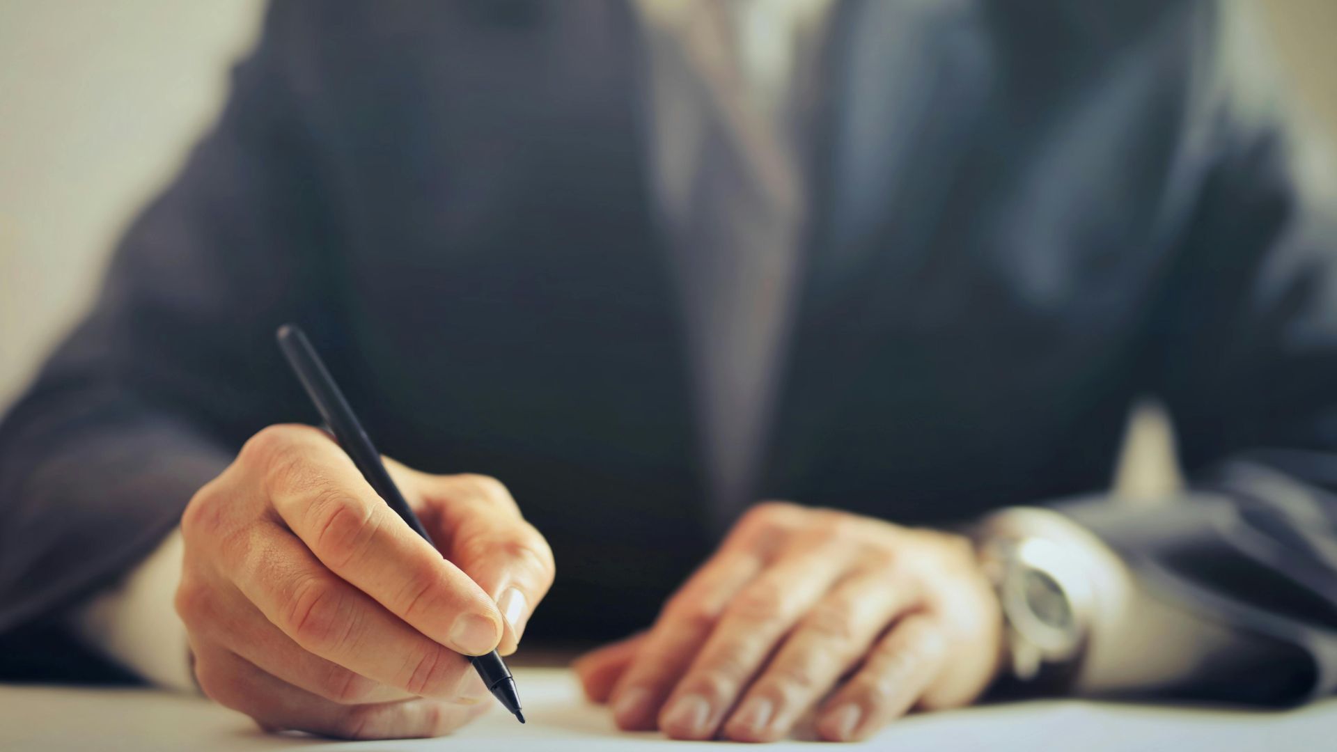 Male entrepreneur wearing formal suit sitting at table in workplace and signing documents while making successful business deal