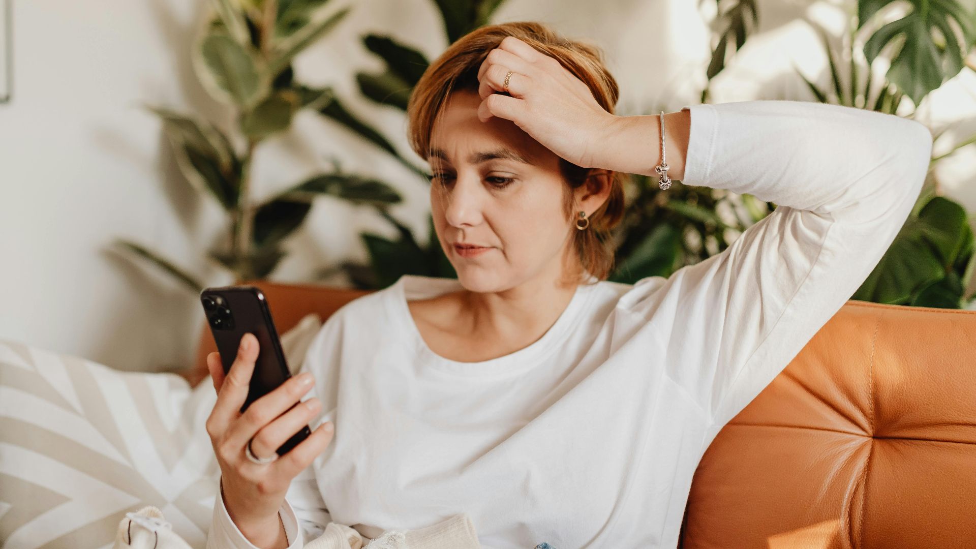 Young woman in white blouse using smartphone while sitting on an orange sofa surrounded by plants.