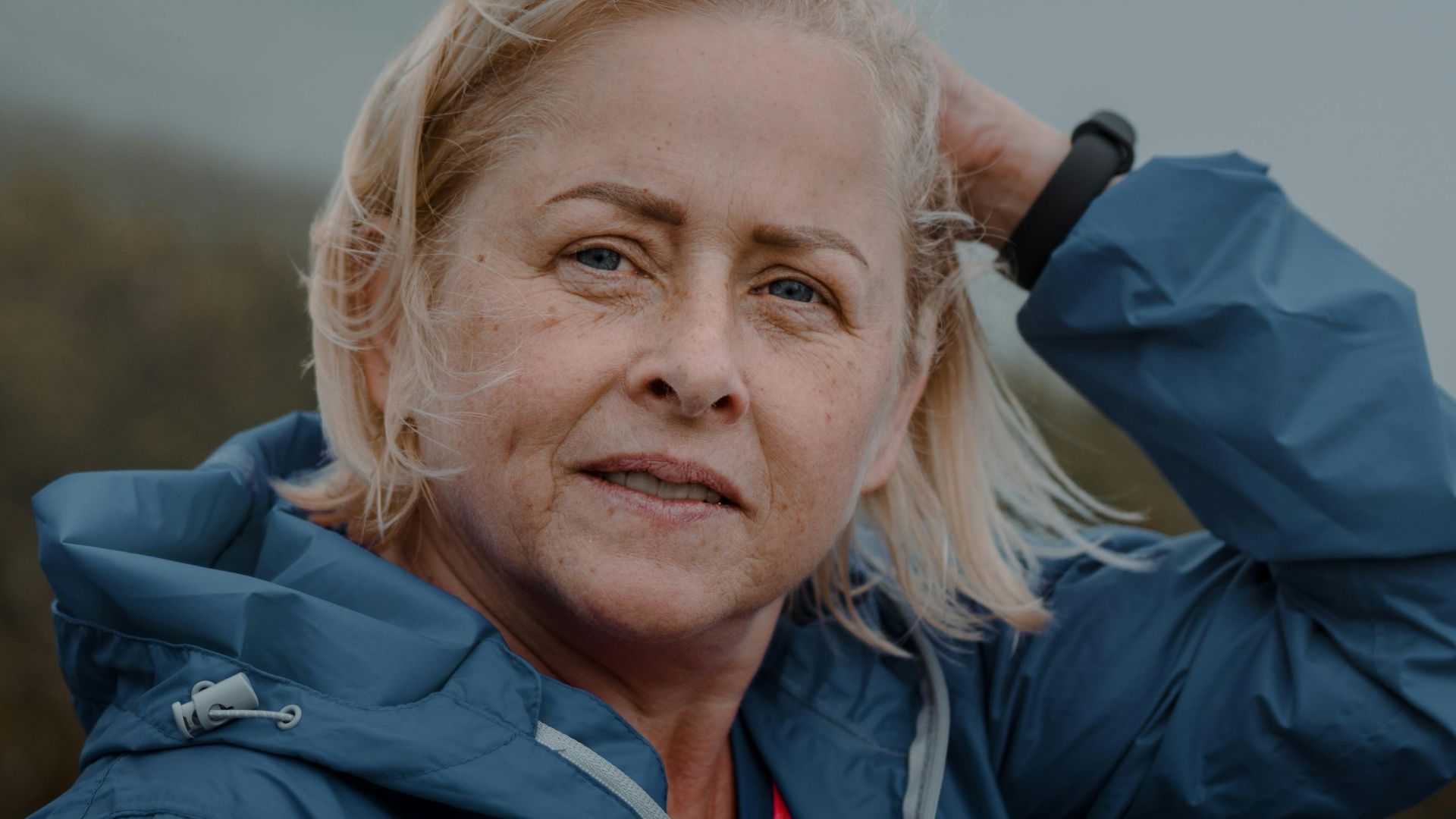 A senior woman outdoors wearing a blue jacket, looking contemplative under a cloudy sky.