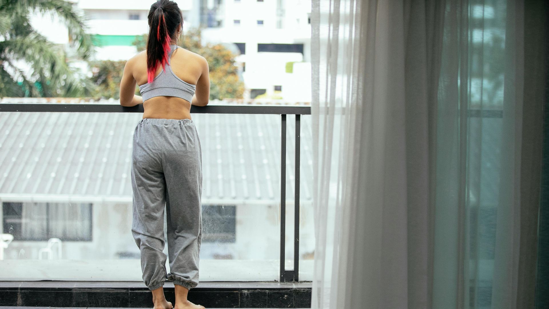 Back view of anonymous peaceful female in activewear standing on terrace with mat and cup of coffee while enjoying morning and observing city