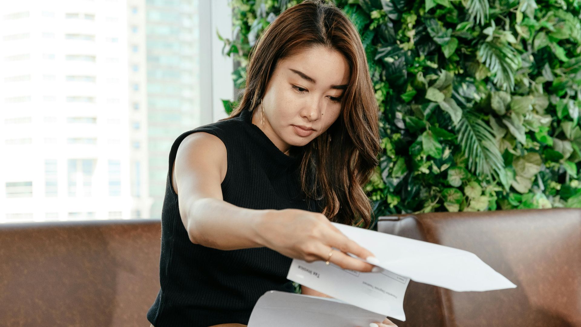 Focused Asian female turning pages of document while sitting on sofa during paperwork in modern workspace with green deciduous plant
