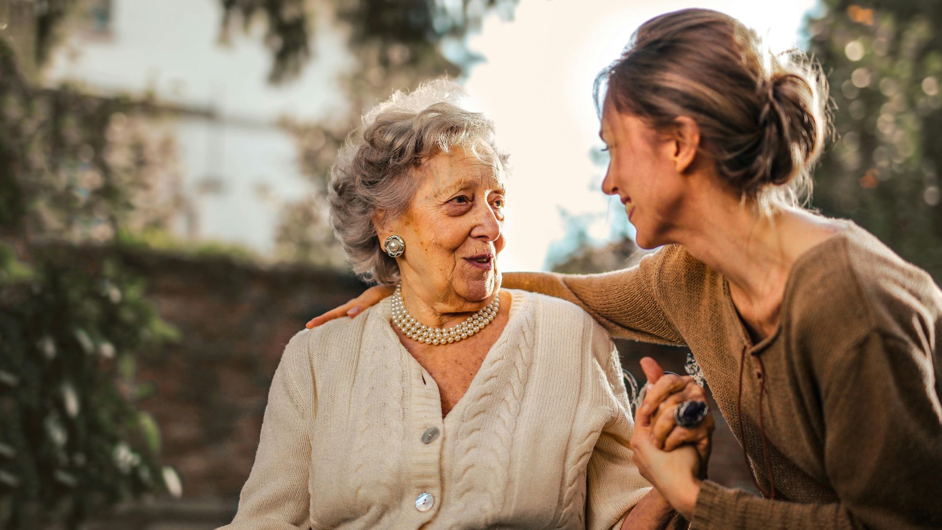 Low angle of cheerful aged lady and adult woman in casual clothes holding hands and smiling at each other while having meeting on yard against blurred green trees and facade of country house in suburb
