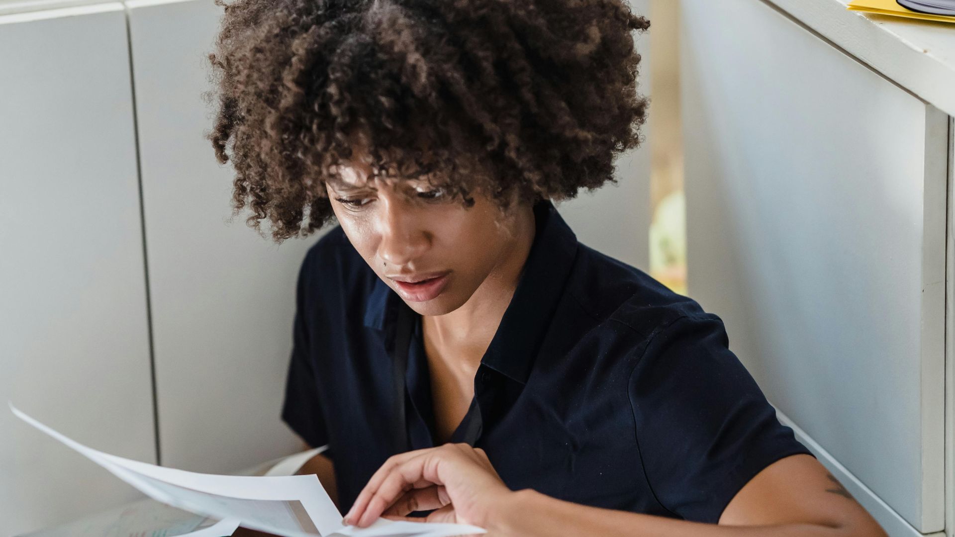 A stressed businesswoman sits on the office floor, reviewing documents with a worried expression.