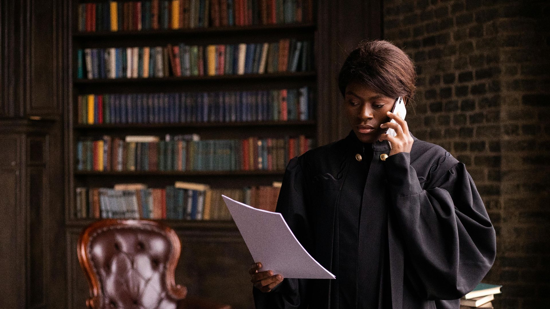 African American judge in traditional robes holds a document and talks on the phone in a law library.