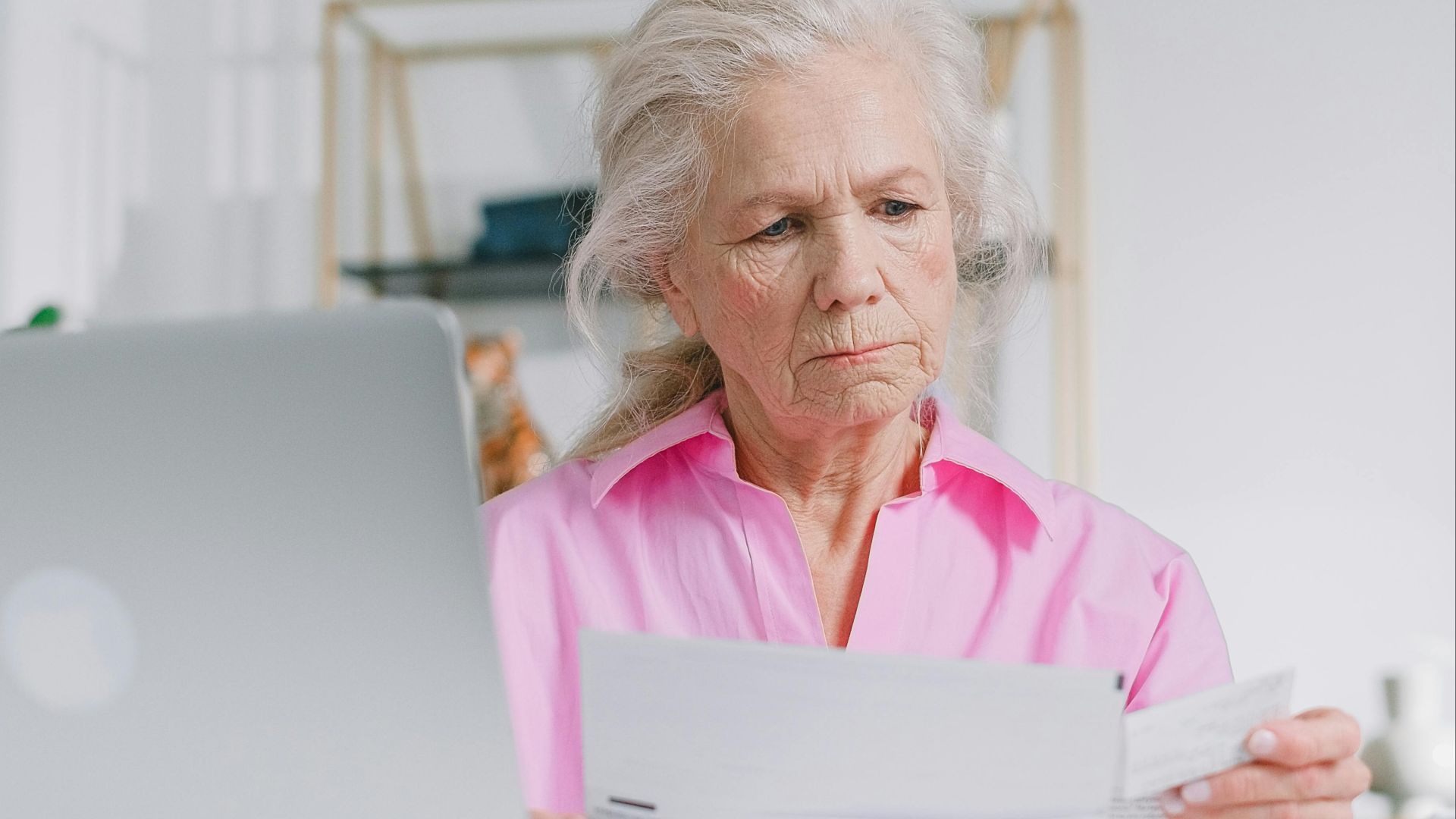 Elderly woman in pink blouse reviewing documents at home office desk.