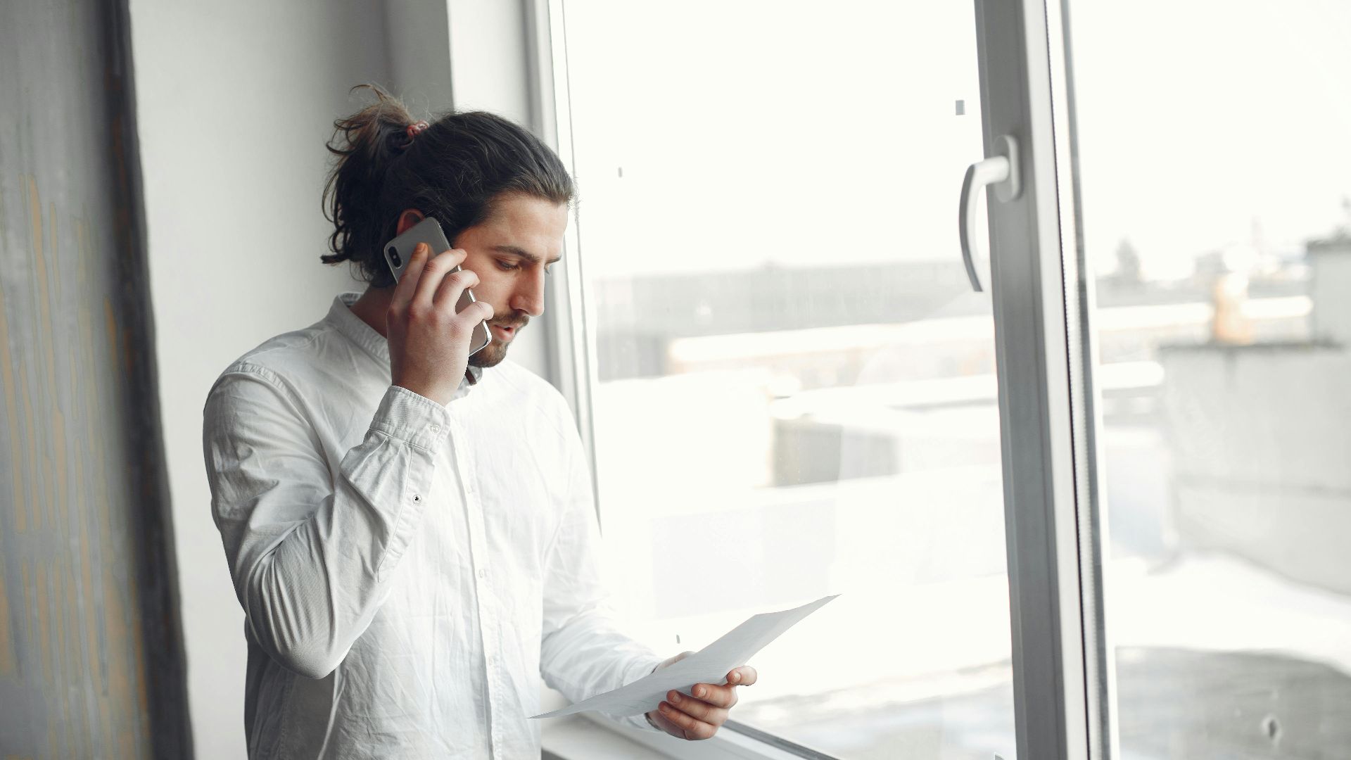 Young man in white shirt, on phone call holding a document, standing by a large window.