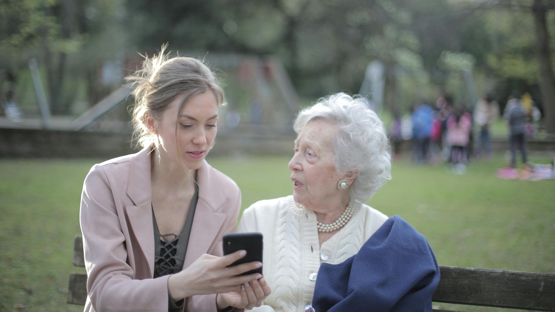 Concentrated female in elegant wear sitting on bench in park and instructing elderly mom on use of cellphone