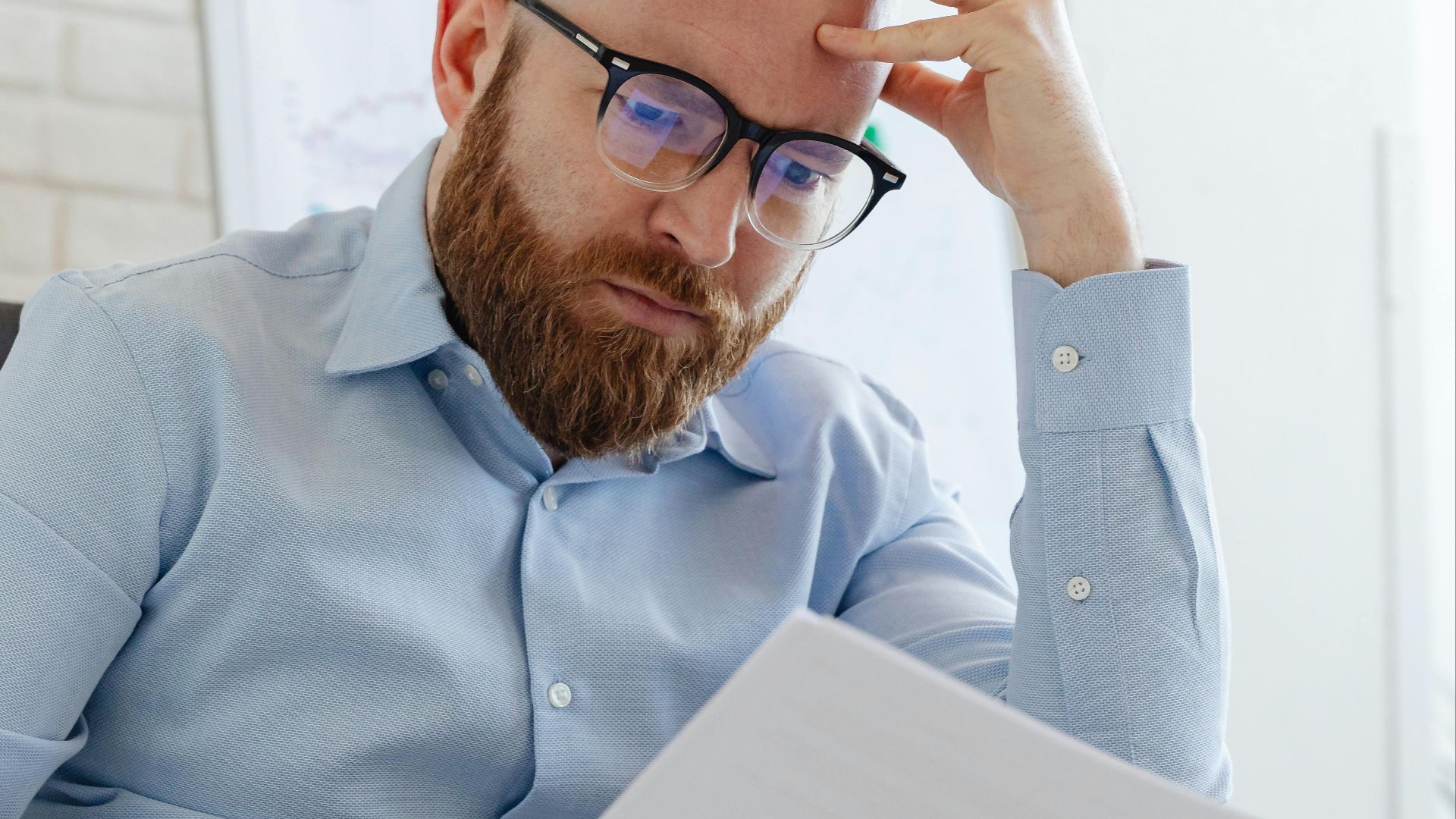 Caucasian businessman with beard and glasses reviewing documents in a modern office setting.