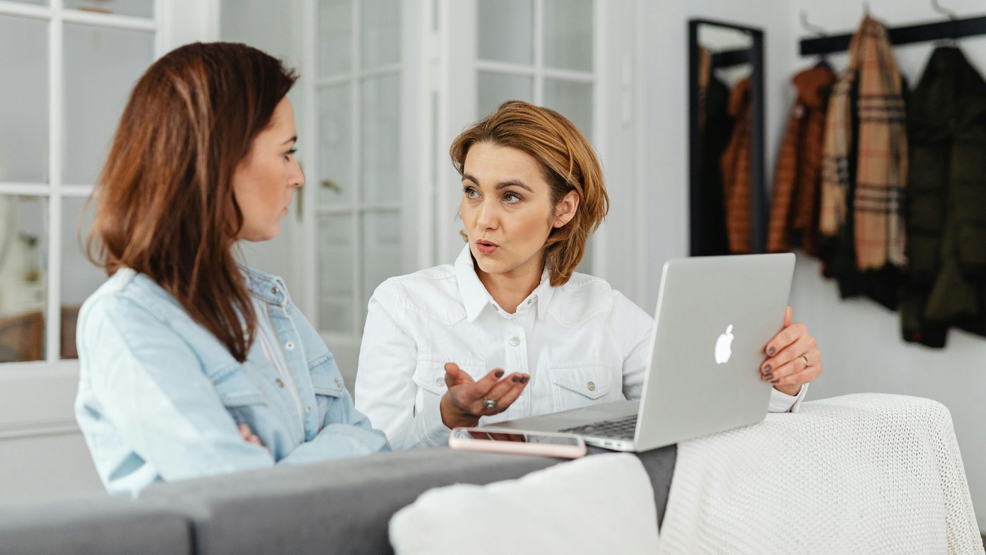 Two women having a conversation while working on a laptop in a cozy indoor setting.