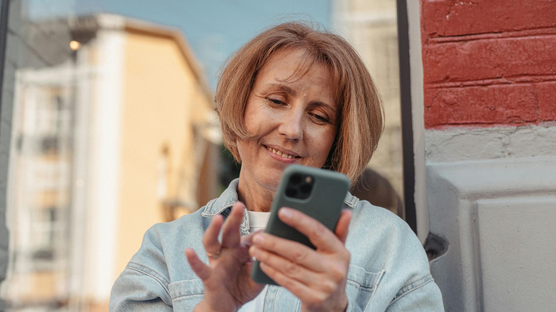 A middle-aged woman in a denim jacket smiles while using her smartphone outside.