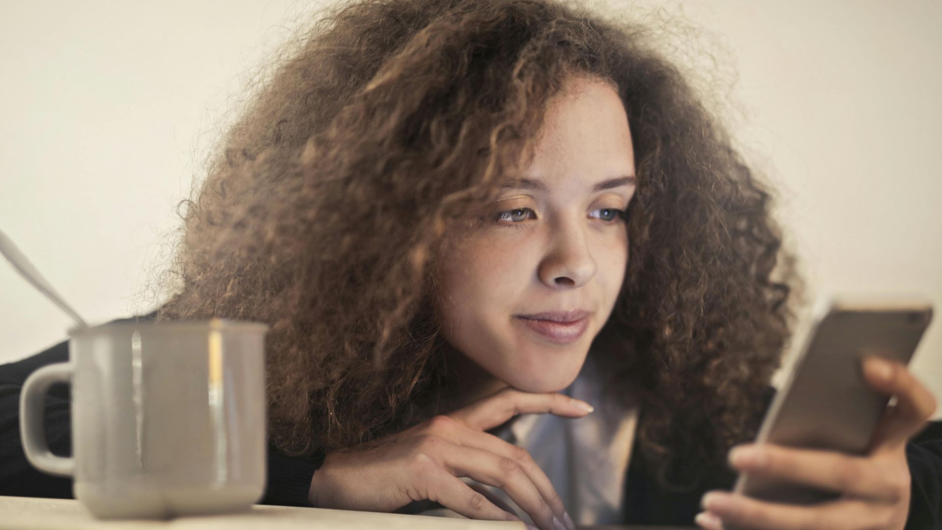 Young woman enjoying a relaxing moment while checking her smartphone indoors with a cup of coffee.