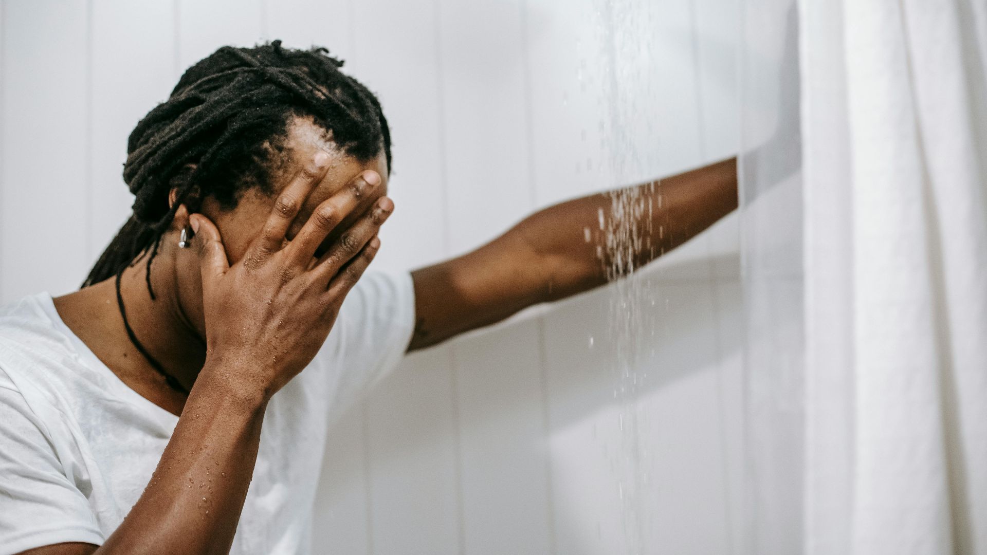 Side view of unrecognizable despaired ethnic male with dreadlocks in white t shirt covering face with hand while standing under shower in bathroom