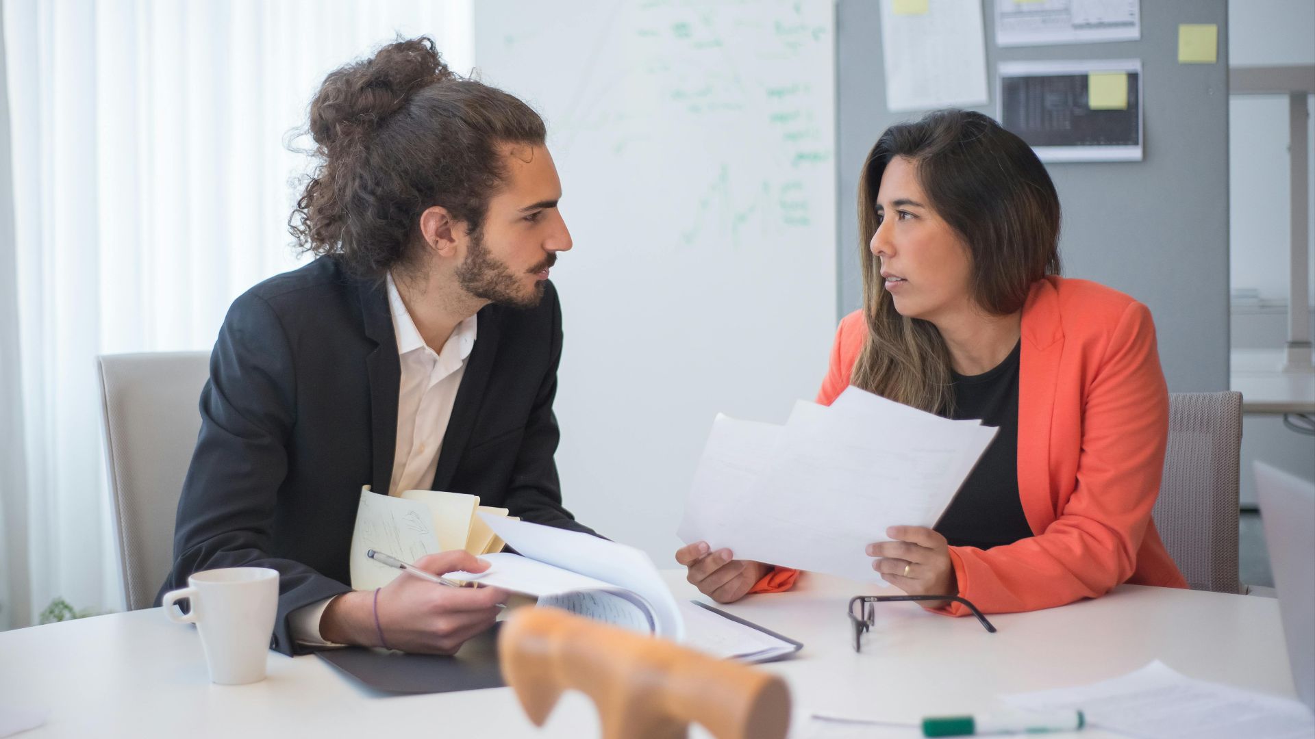 Two colleagues engaged in a discussion at an office table with documents and coffee.