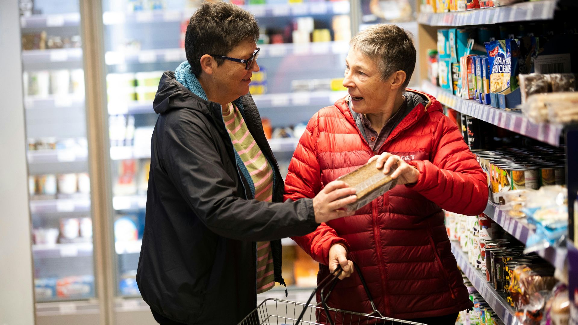 Two elderly women interact joyfully while shopping in a supermarket aisle.