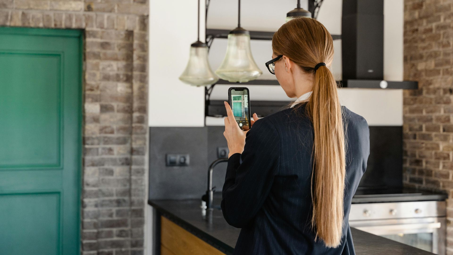 Rear view of a woman with long hair taking a photo with a smartphone in a stylish kitchen.