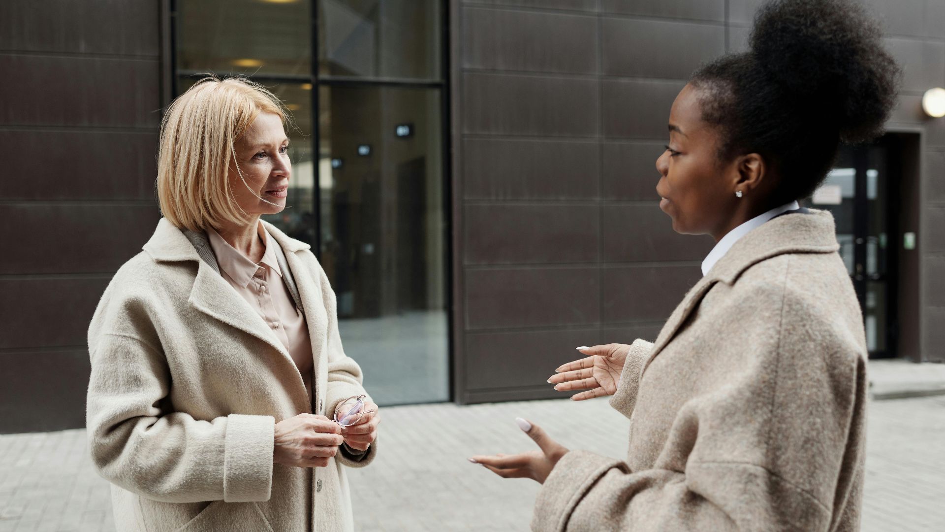 Two businesswomen in coats converse outside a modern office building.