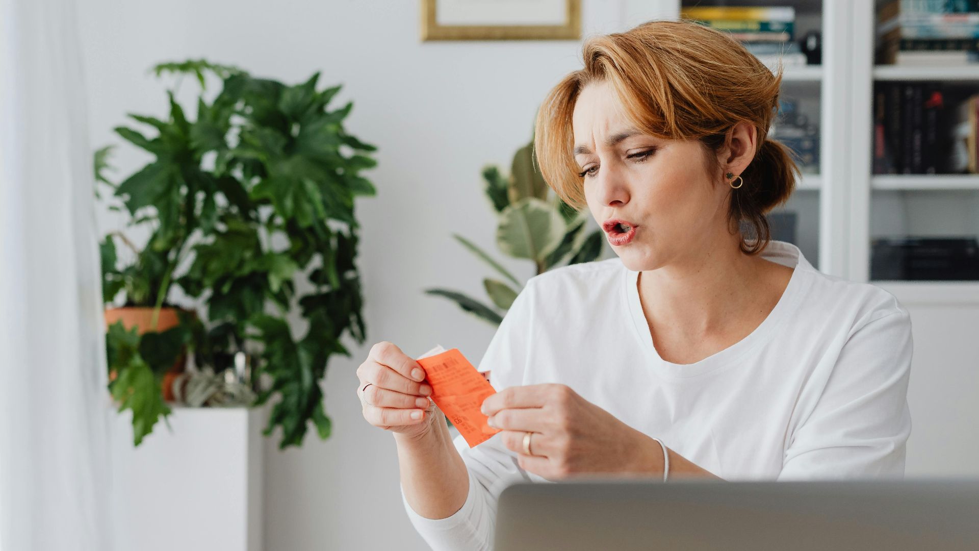 Woman reviewing bills at home desk with laptop and plants, managing personal finances.