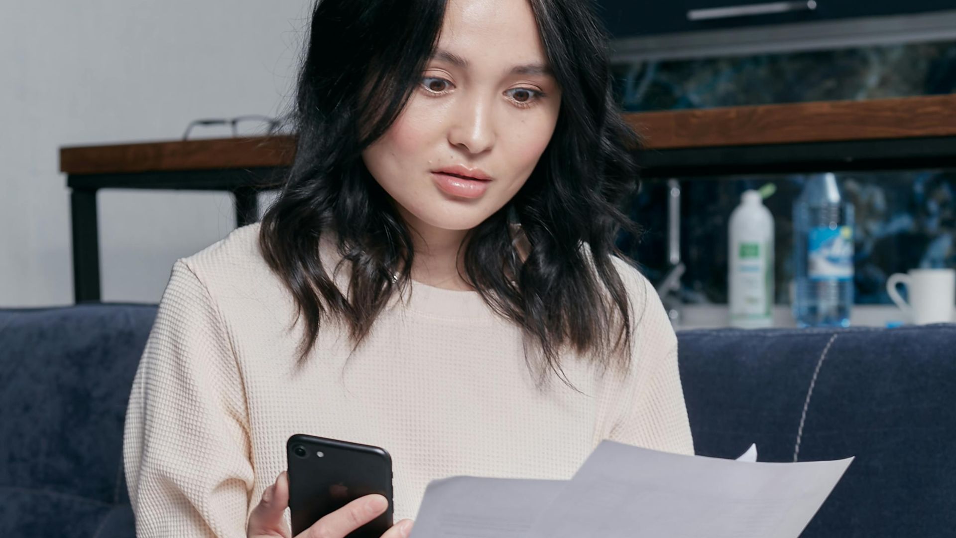 Young woman reviews documents and phone indoors, showcasing concern while seated on a couch with scattered papers. Business concept.