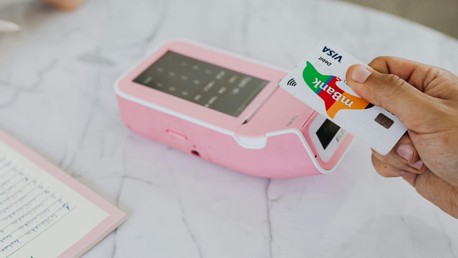 A hand using a Visa card for a contactless payment on a pink terminal.