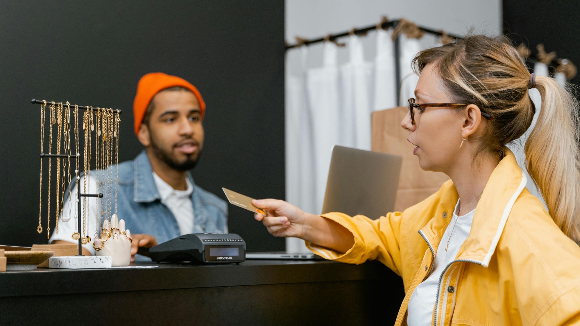 Woman in yellow jacket pays with credit card at shop counter with jewelry display.