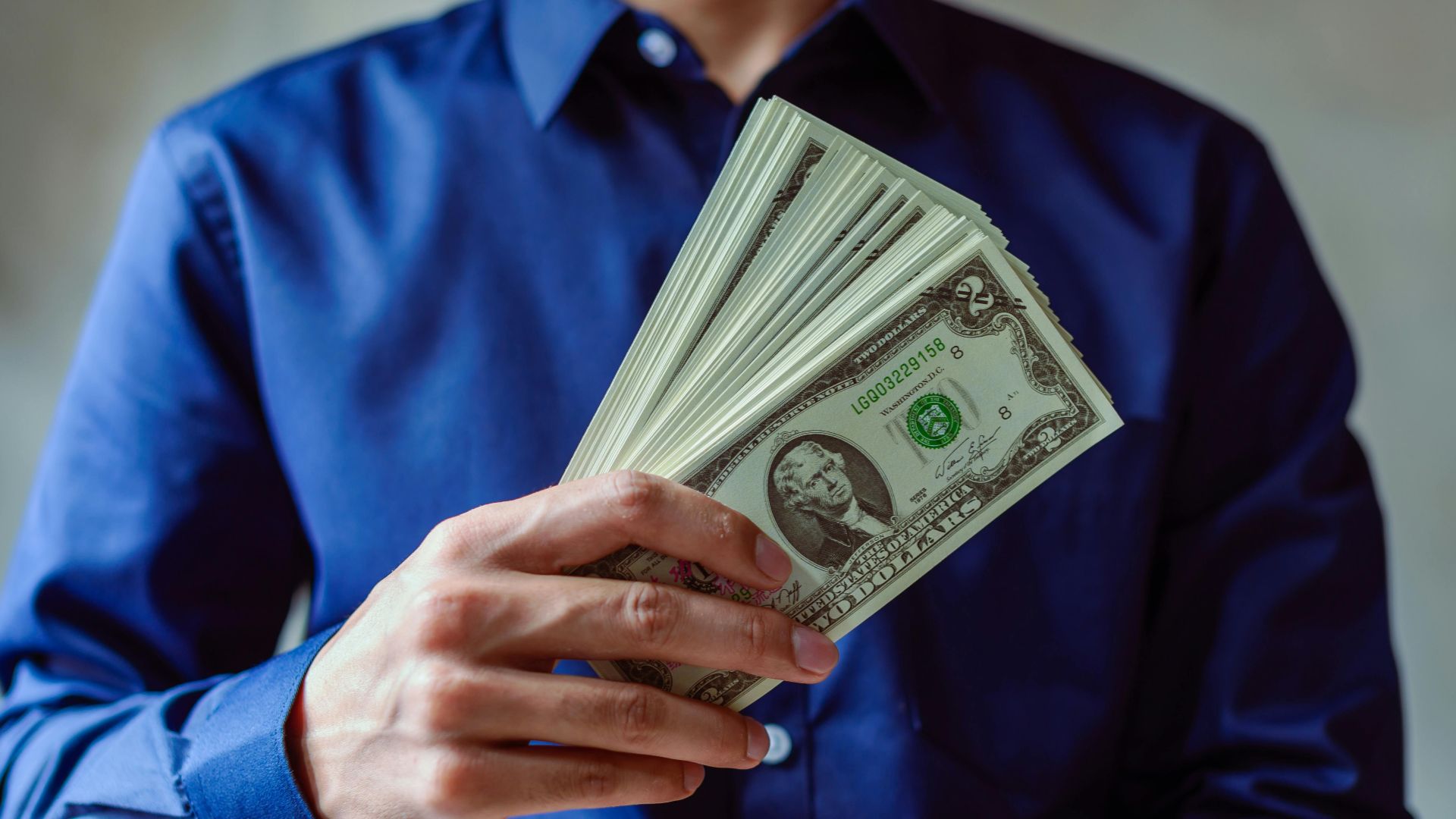Close-up of a person in blue shirt holding multiple US dollar bills.