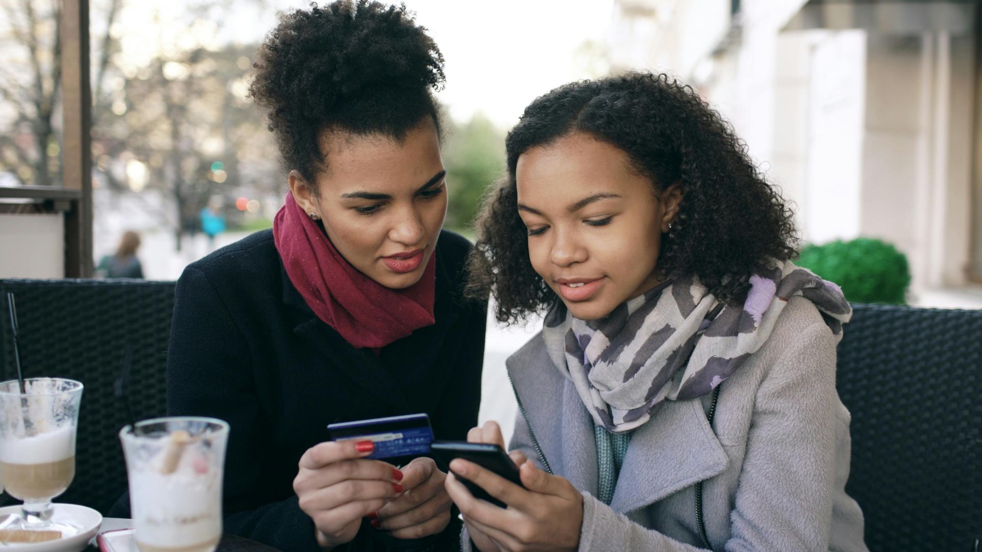 Two women enjoy online shopping using a smartphone and credit card at a cozy outdoor café.