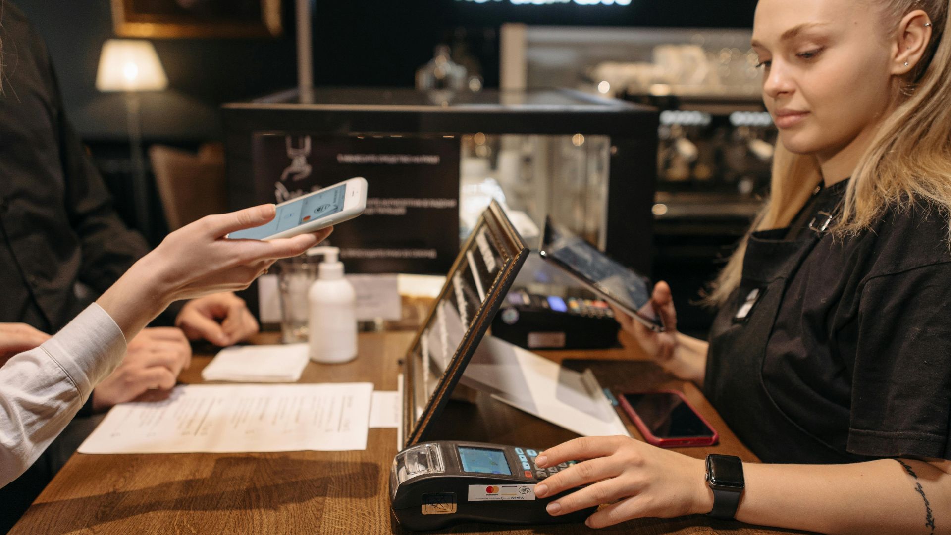 Customer using mobile phone for contactless payment at a coffee shop counter with a barista.