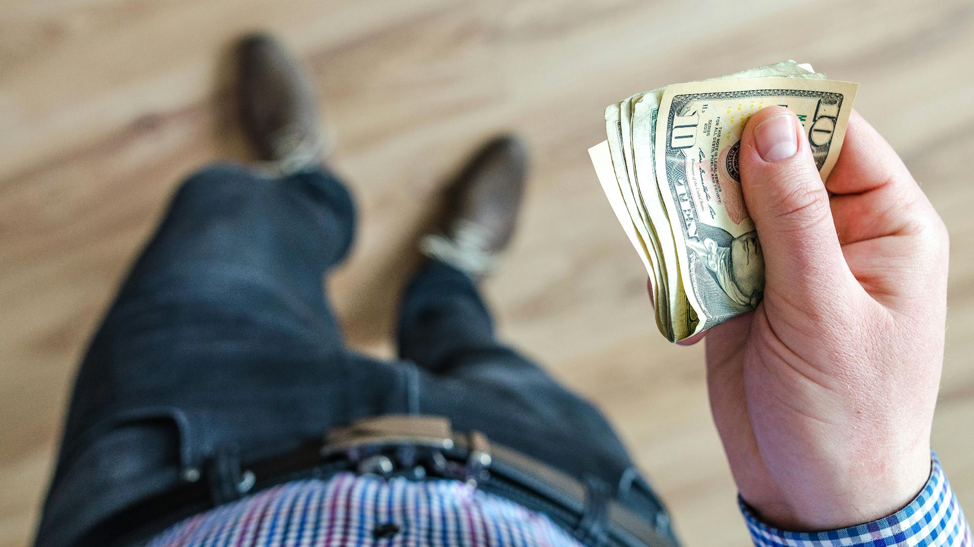 Close-up shot of a man holding US dollar bills indoors with a wooden floor background.