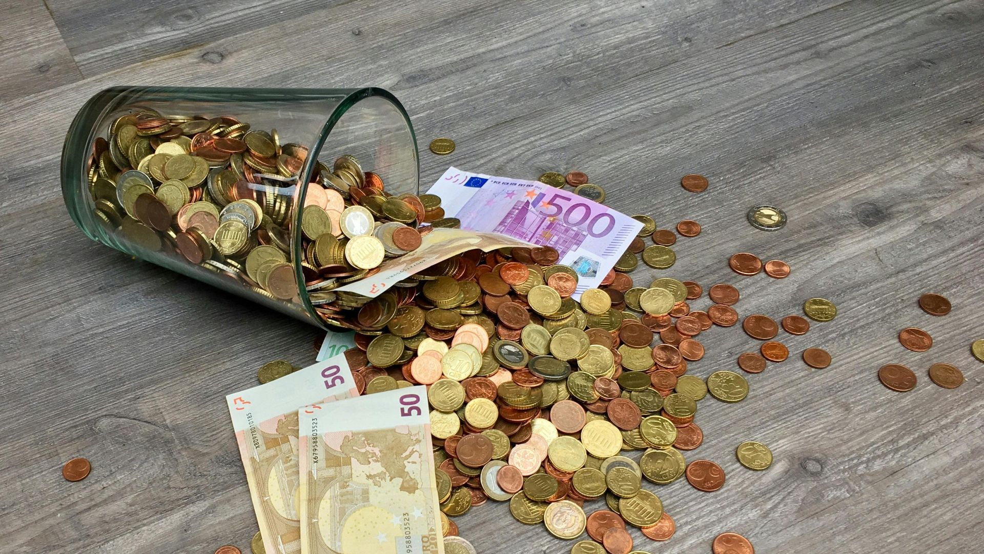 A glass jar with euro coins and banknotes spilled on a wooden surface, representing finance and savings.