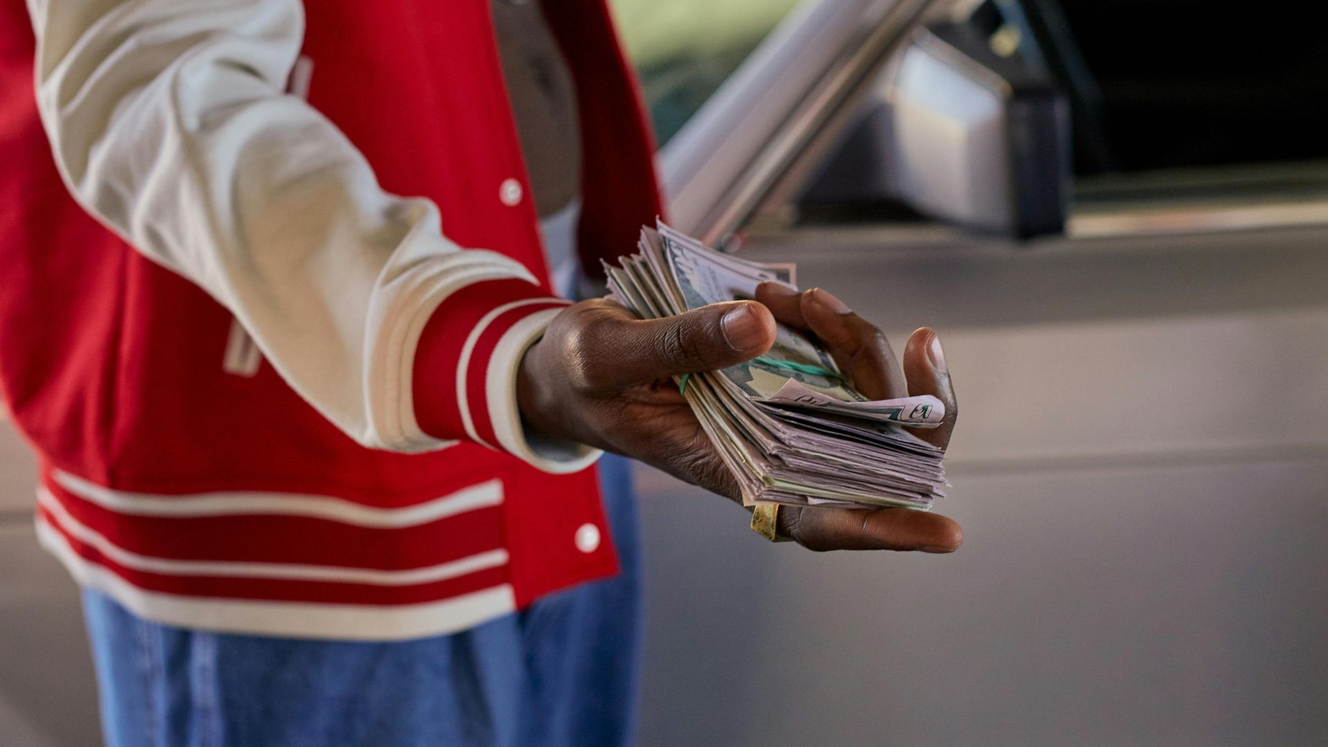 Close-up of a person offering a stack of cash in front of a car, symbolizes financial transaction.