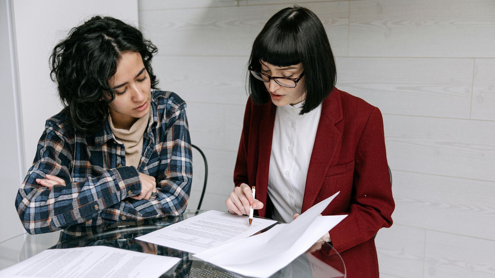 Two women collaborating at a glass table, reviewing business documents in a modern office setting.