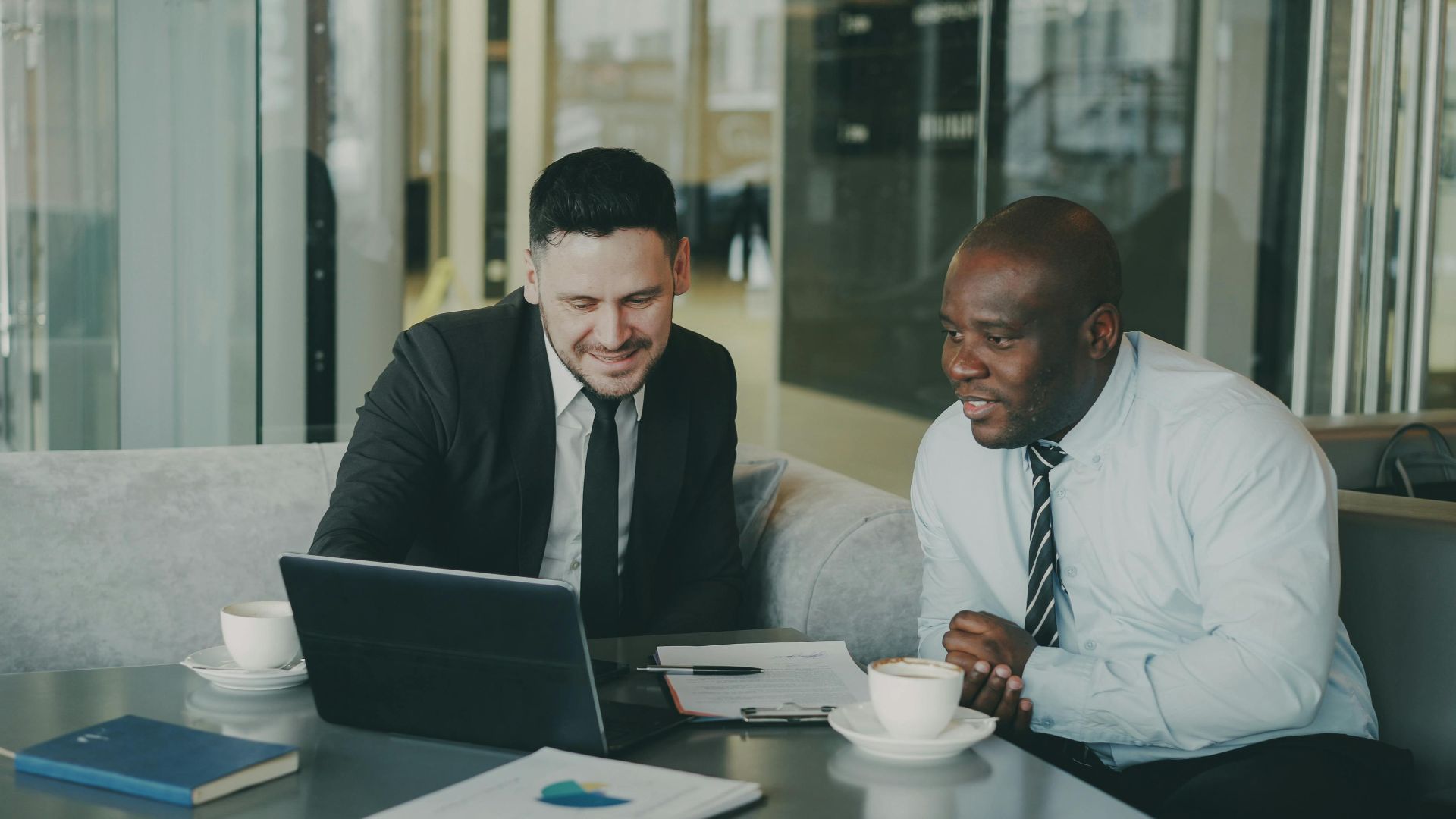 Two businessmen discussing work on a laptop in an office with coffee cups nearby.