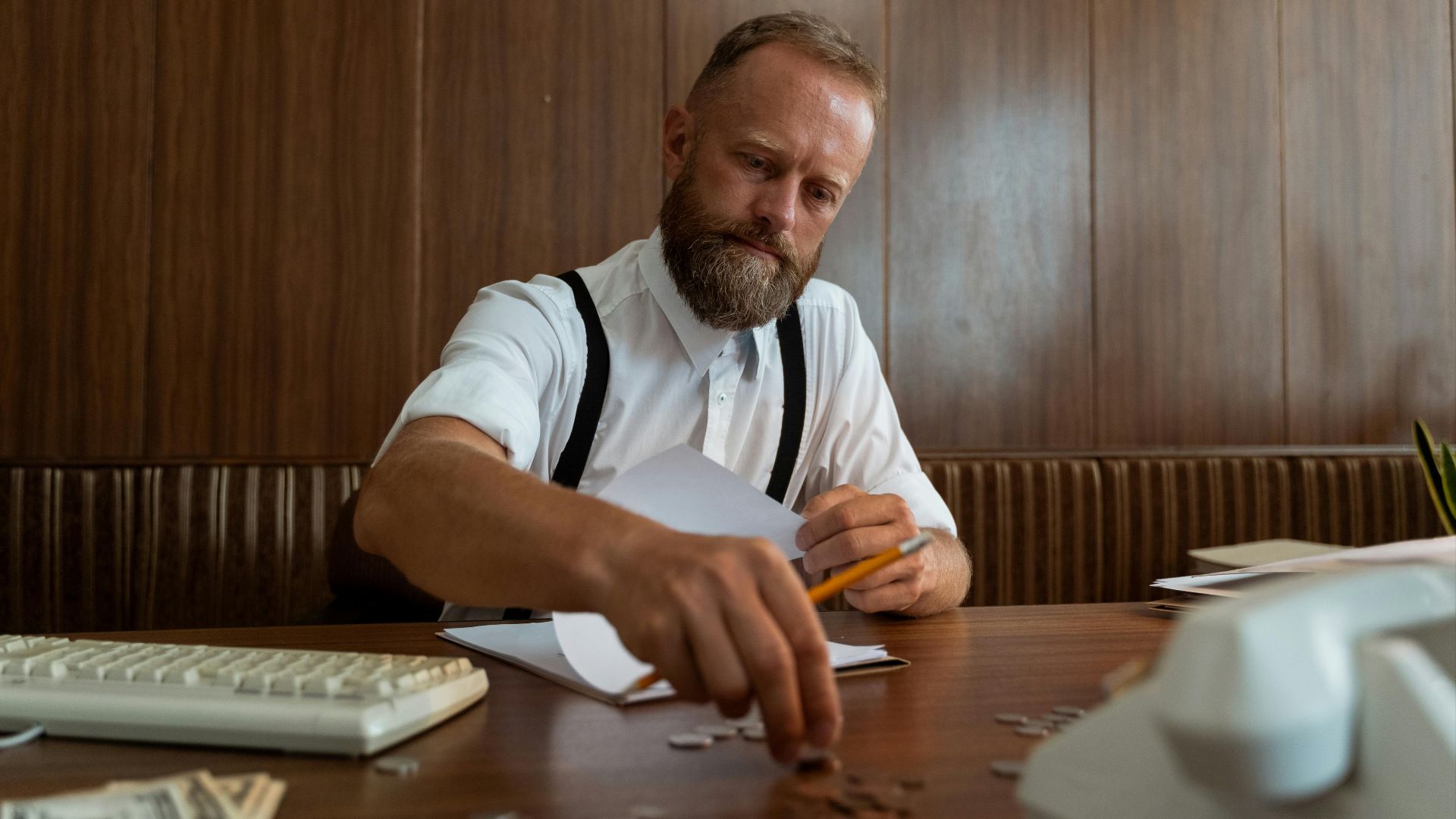 A bearded businessman wearing suspenders counts coins at a vintage workspace, embodying retro office style.