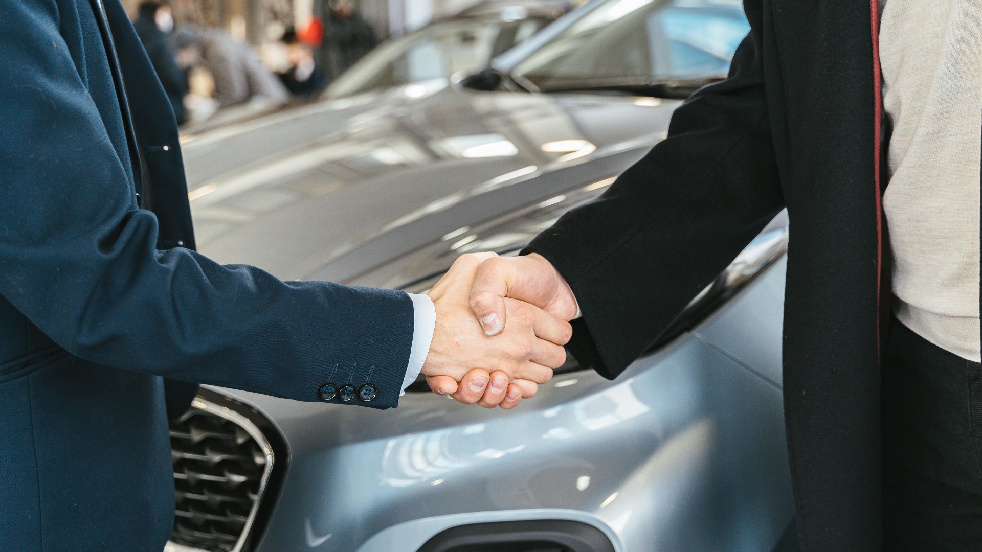 A close-up of two people shaking hands in front of a car in an indoor setting.