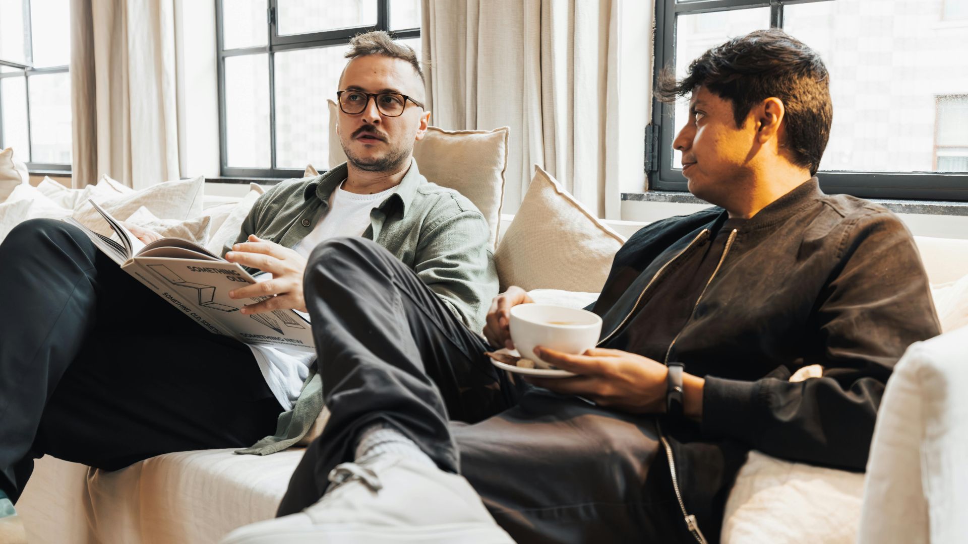 Two men sitting on a cozy sofa, one reading a magazine, enjoying coffee in a bright living room.