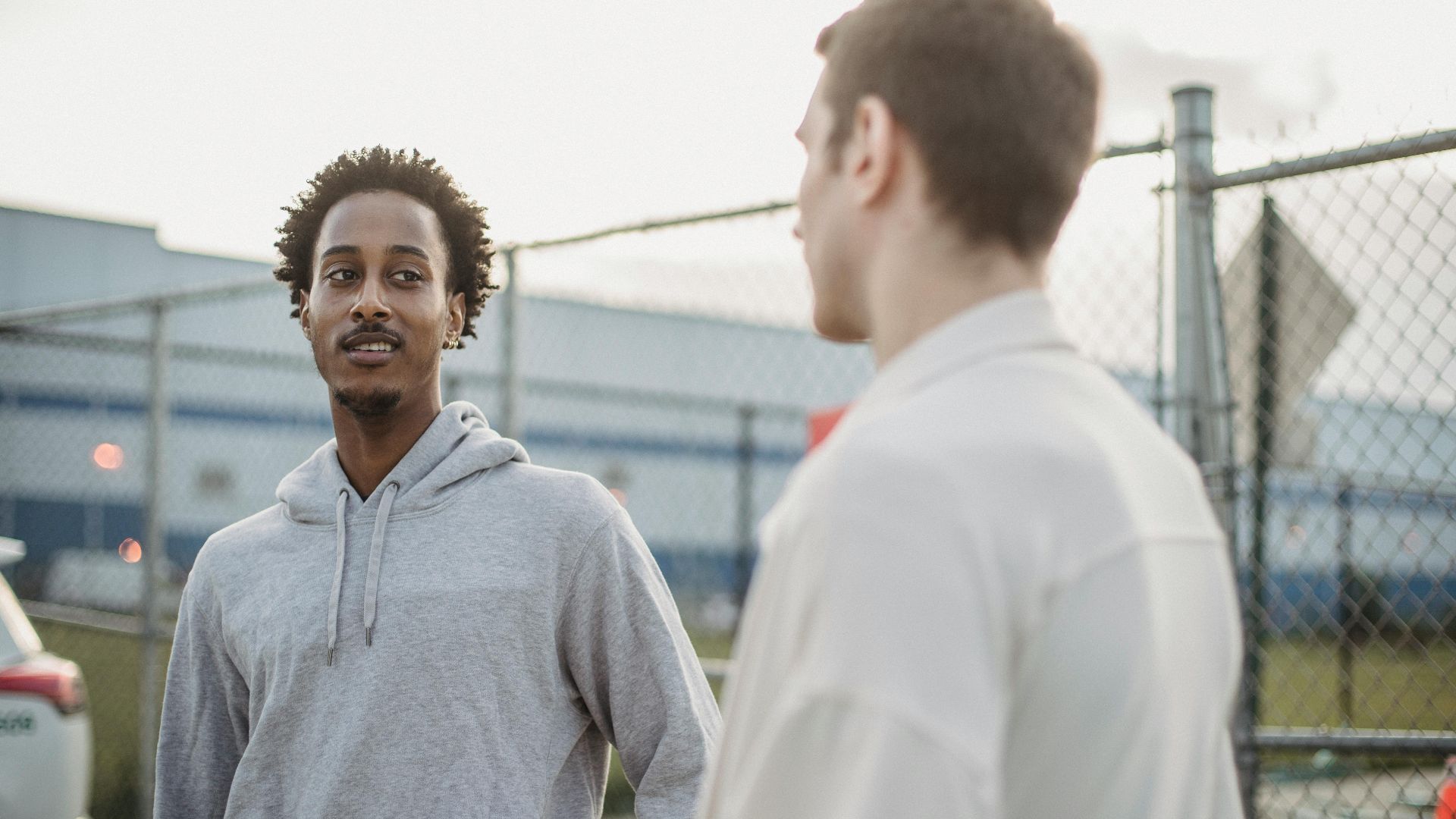 African American male standing with friend against metal grid near car on parking lot