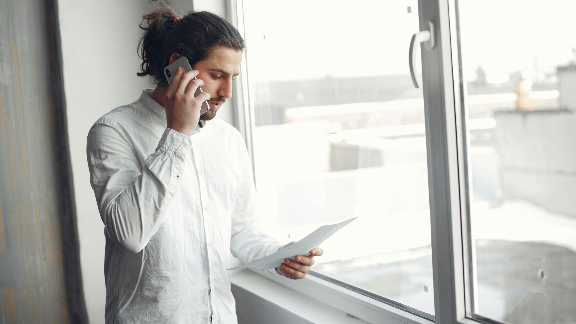 Young man in white shirt, on phone call holding a document, standing by a large window.