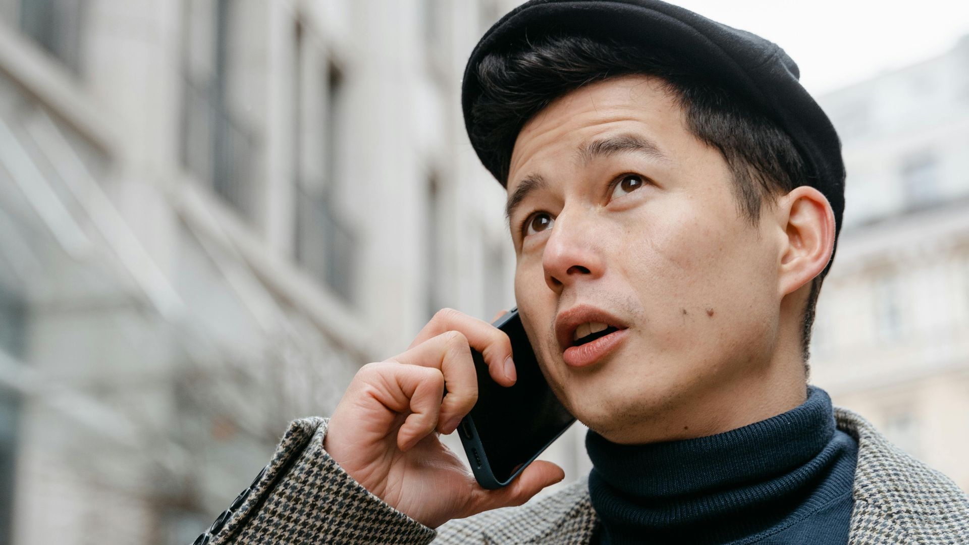 Stylish man in a city talking on a smartphone outdoors, wearing a coat and hat.