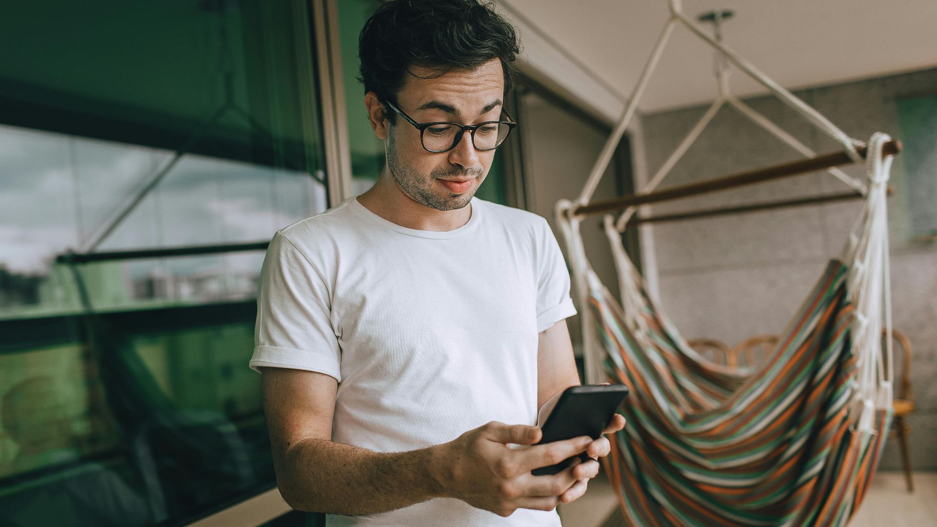 Young man in glasses using smartphone indoors with striped hammock.
