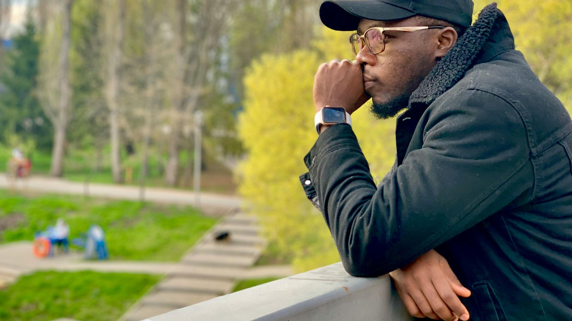 A man leans on a railing, thoughtfully gazing at a park in springtime, wearing a cap and jacket.