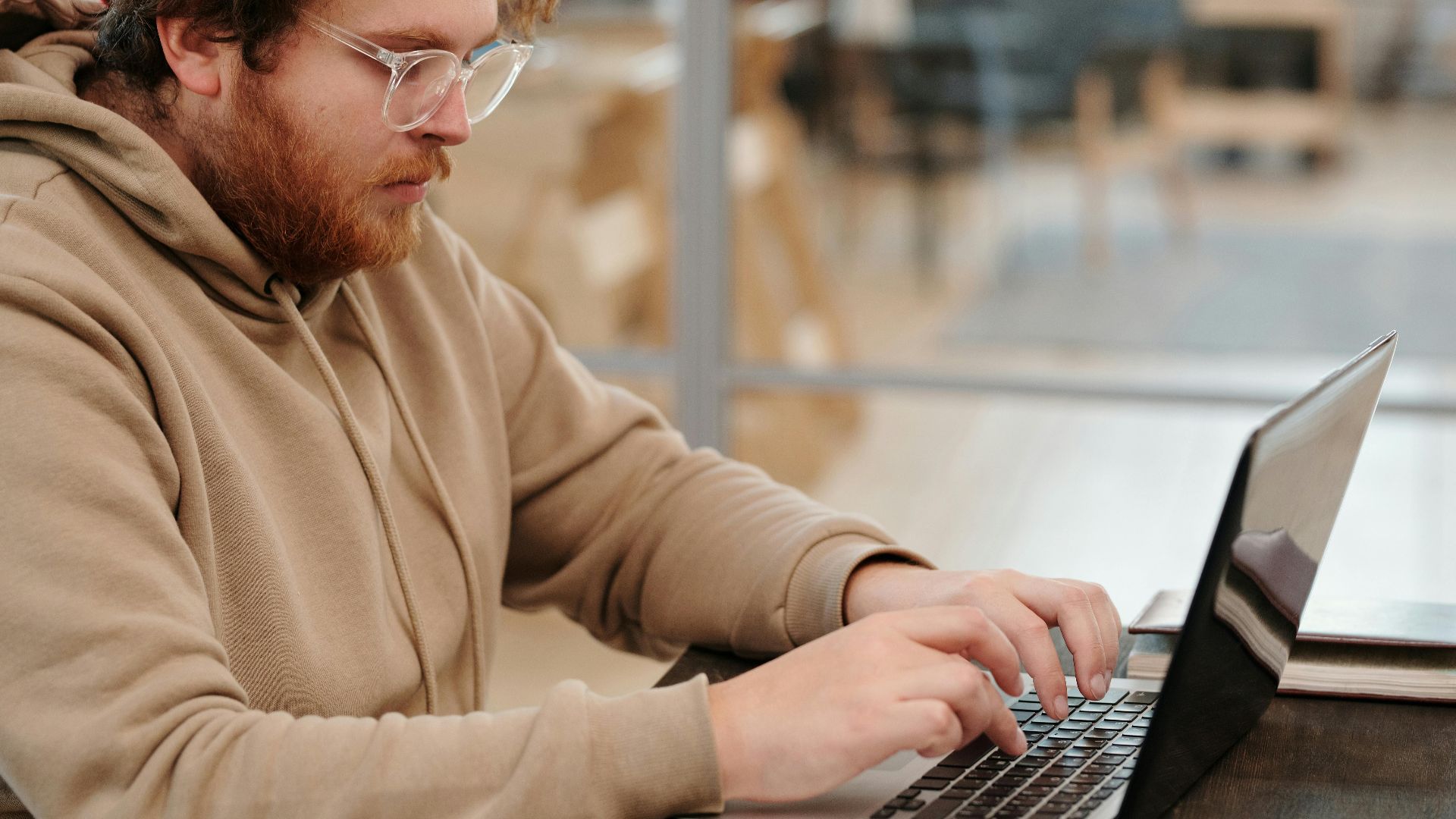A bearded man wearing glasses, working intently on a laptop in a cafe setting.