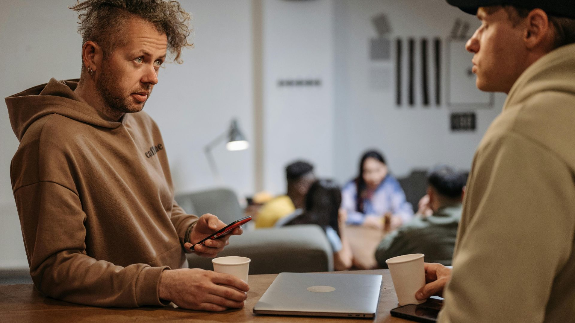 Two men in a casual office setting talking over coffee with laptops.