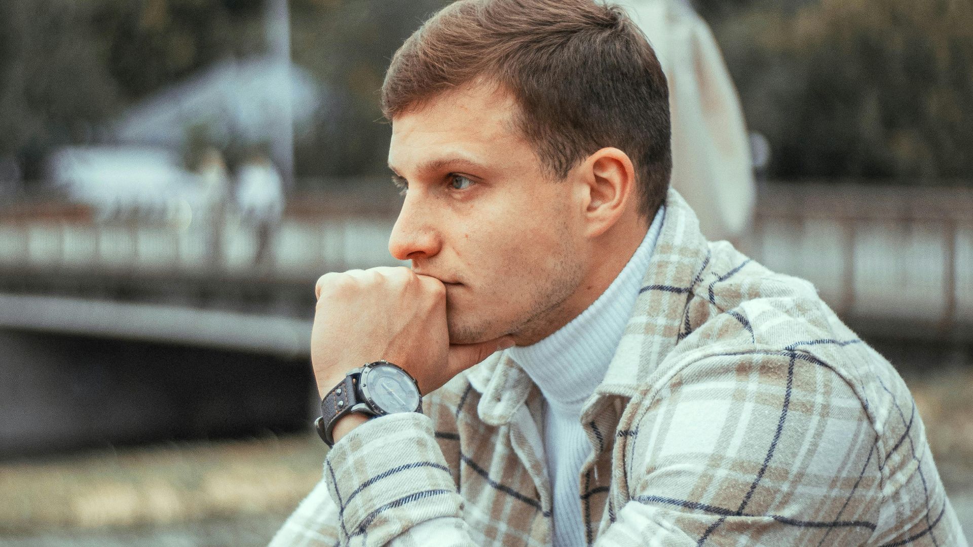 Close-up of a man in a plaid jacket, deep in thought at an outdoor cafe in a city.